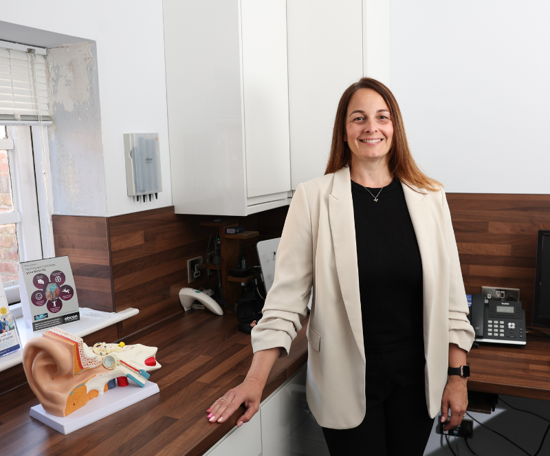 Audiologist Kim at House of Hearing Knutsford, standing in front of a desk with a model of the human ear.