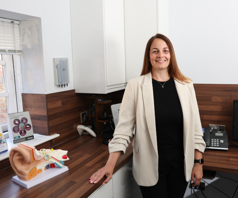 Audiologist Kim at House of Hearing Knutsford, standing in front of a desk with a model of the human ear.